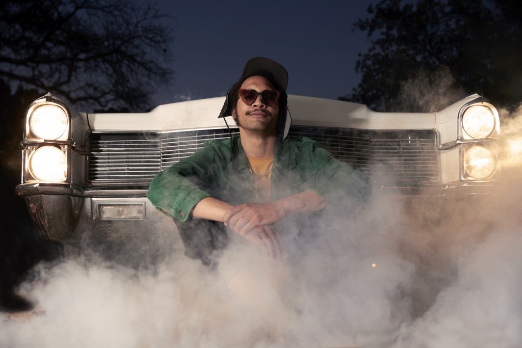 A promotional image for artist Tré Burt. Burt is pictured sitting in front of a car, the car is smoking and Burt is wearing sunglasses, a jacket, and a hat. 