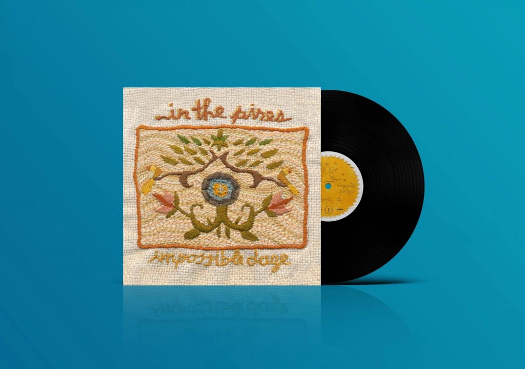 An image of In the Pines' record "Impossible Daze." This is a promotional image, with an image of the record on vinyl, half outside of the sleeve, which has a brown and rust colored embroidered design made up of flowers. The image is against a blue background.