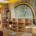 A child care room has a rainbow on the wall with a pot of gold. There are cubbies to keep things up against the same wall. The tables nearby are well organized with cubs of coloring materials in them.
