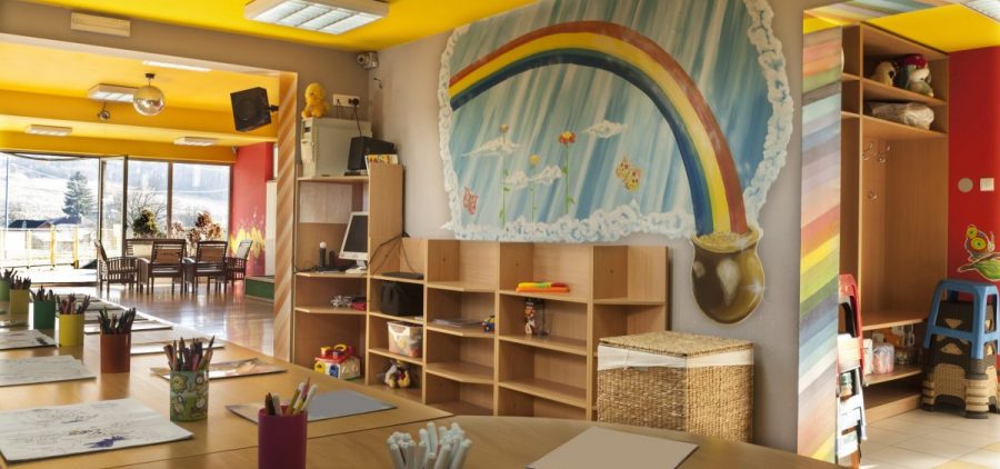 A child care room has a rainbow on the wall with a pot of gold. There are cubbies to keep things up against the same wall. The tables nearby are well organized with cubs of coloring materials in them.