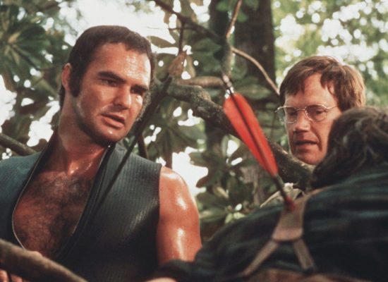 A still taken from the movie "Deliverance," with two people in the shot, examining the spot where an arrow has stuck into a tree. They are in a wooded area.