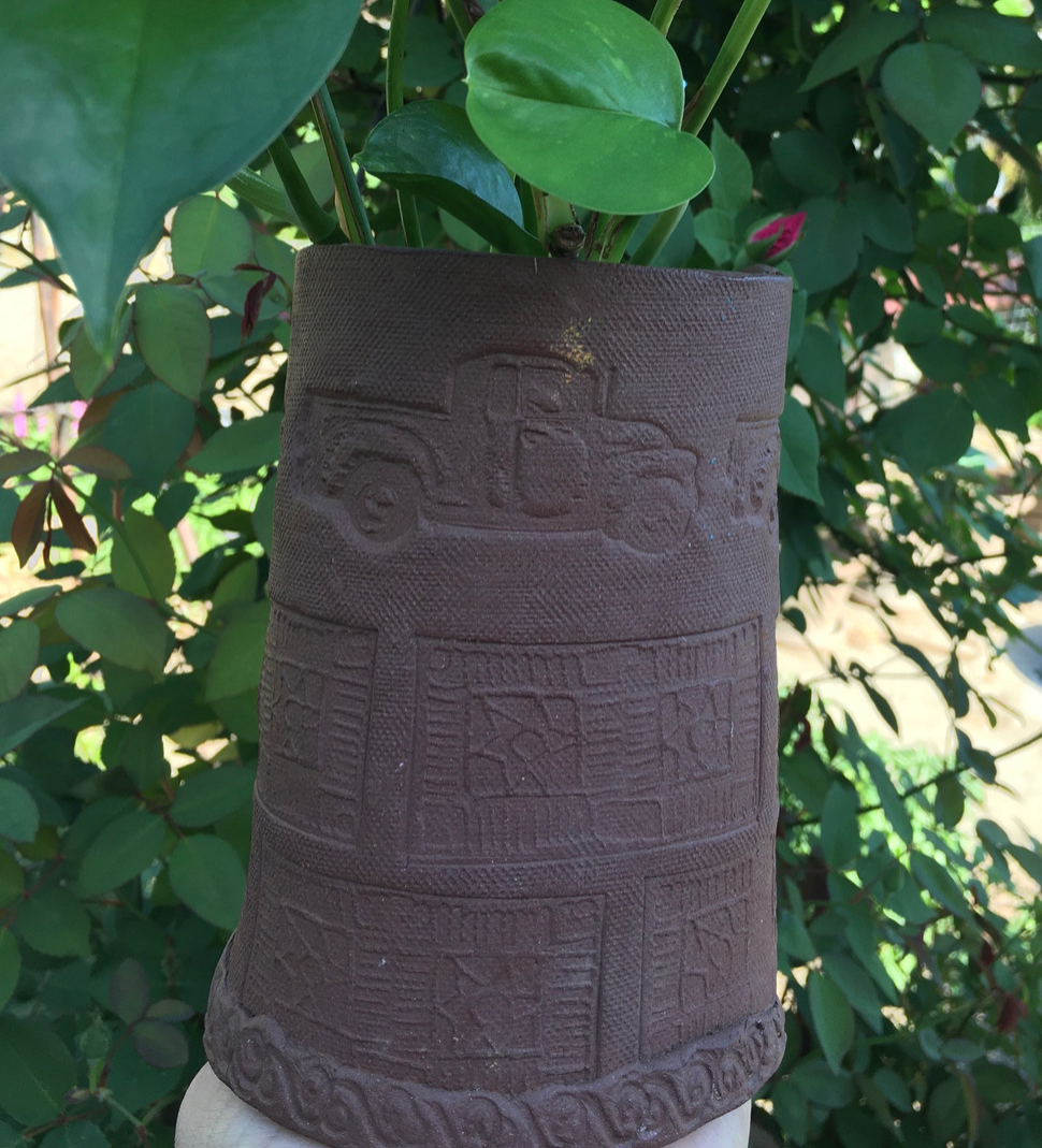 A cylindrical garden pot that is textured elaborately, and has a green plant planted in it. The background is the interior of a wooded area.