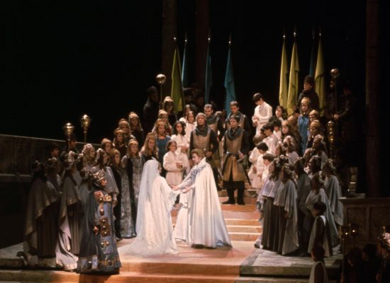 A promotional image for Wagner’s Lohengrin - two people in flowing white robes are surrounded by people dressed in dark clothing.