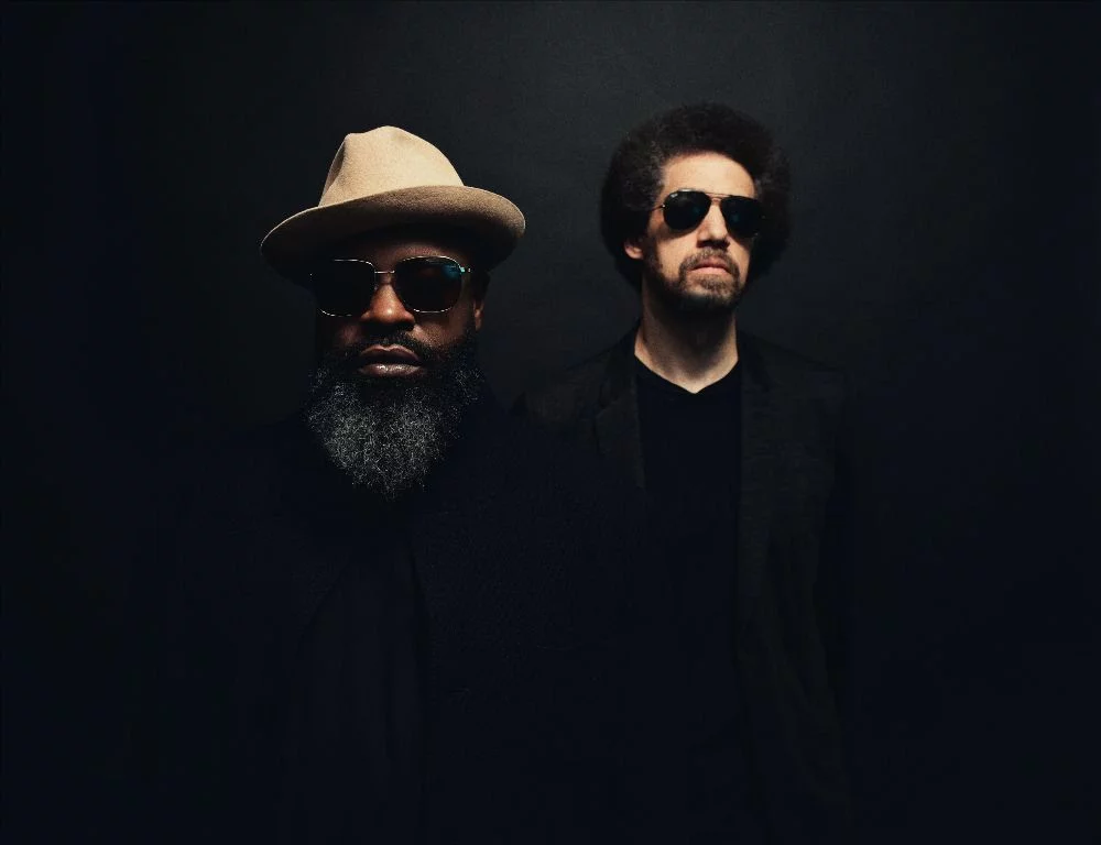 A promotional image for Tarik Luqmaan Trotter, who performs as Black Thought. Trotter is in the foreground with a hat, shrouded in darkness, while producer Danger Mouse is to his shoulder, wearing sunglasses.
