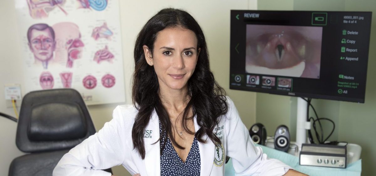 Dr. Yael Bensoussan poses for a photograph in a white doctor's coat with a hand on a machine that analyses vocal chords.