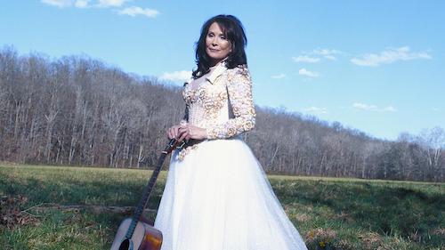 Loretta Lynn in white dress holding guitar in field
