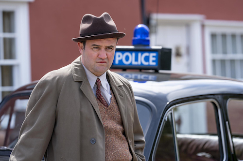 male detective in hat, leaning against police car