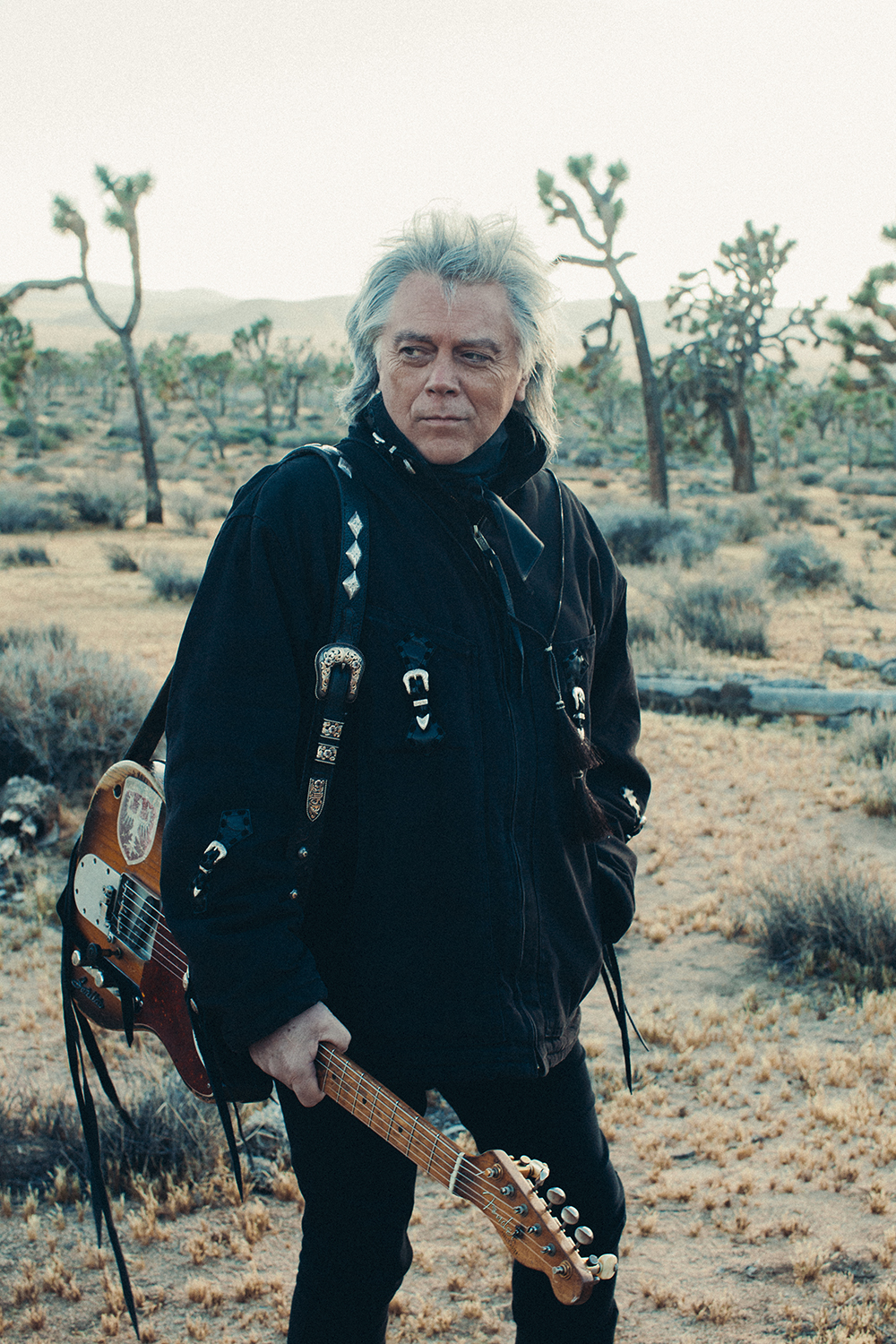 A promotional image of Marty Stuart, he is holding a guitar and is seemingly in a desert