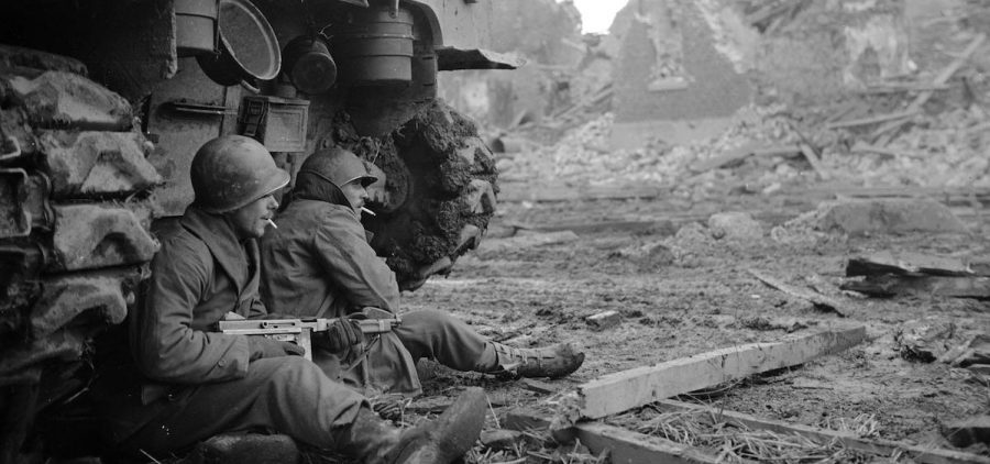 Two WWII soldiers sitting on ground behind tank, rubble in the background