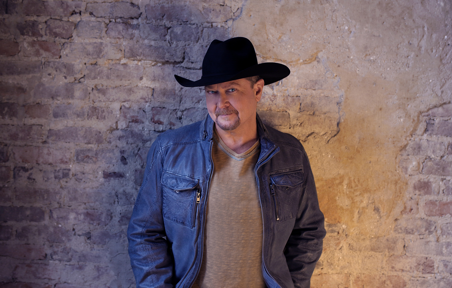 A promotional image of Tracy Lawrence, he is posing against a brick wall and is wearing a cowboy hat.
