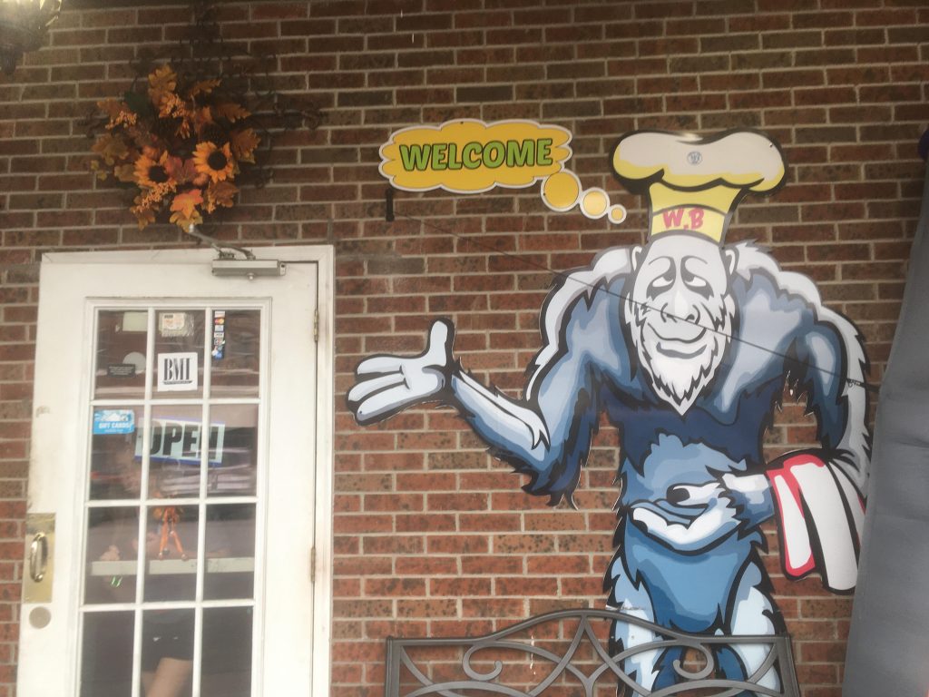 The Wood Booger Bar & Grill has a sasquatch looking thing illustrated on the storefront. It is wearing a chefs hat and has a thought bubble that reads "Welcome"