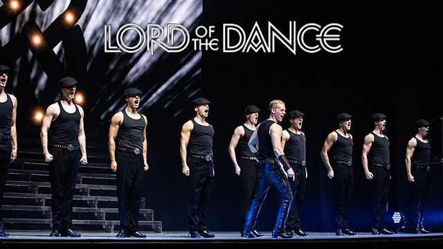 A promotional image from "Michael Flatley's Lord of the Dance." The image shows a line up of men in hats and black tank tops on a stage, ready to dance.