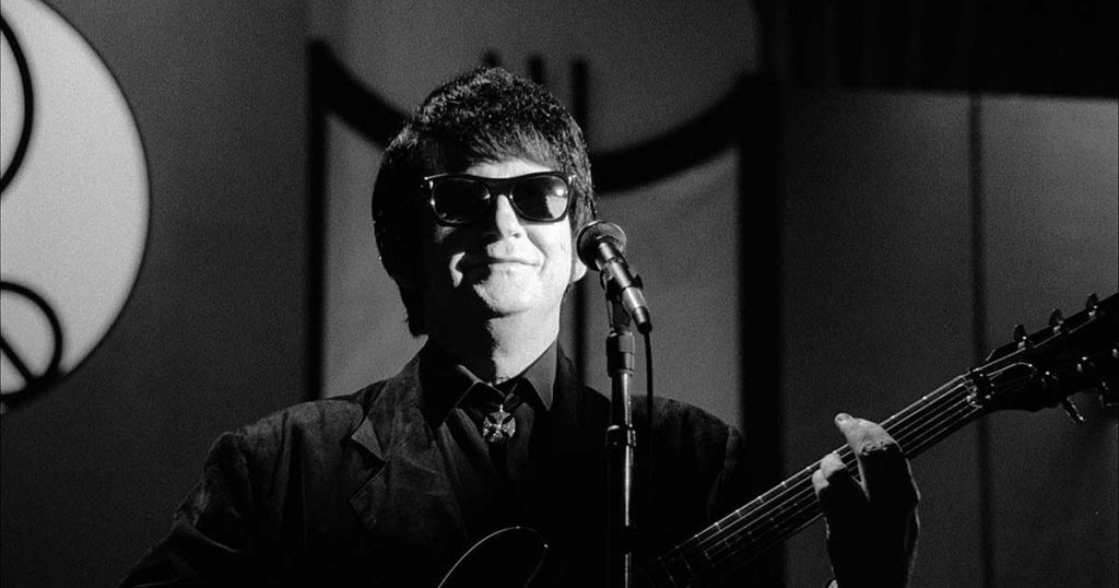 A black and white image of musician Roy Orbison, wearing his iconic sunglasses and holding a guitar.