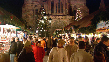 An image from "Rick Steves European Christmas" depicting a Christmas market in Germany. Many people are outside at night under festive lights.