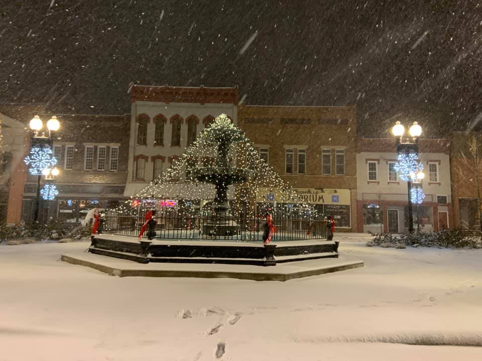 A photograph of the water fountain in downtown Nelsonville during the wintertime at night. It is fetsively decorated and it is snowing.