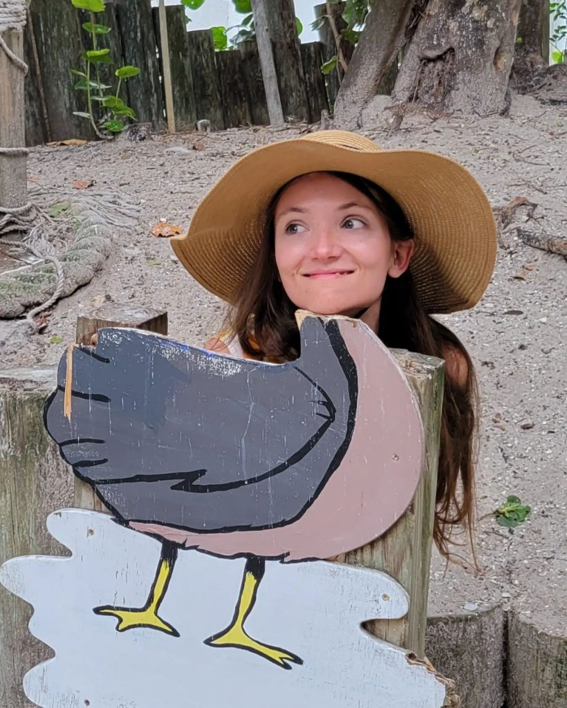 An image of Emily Votaw wearing a wide brimmed straw hat and posing in front of a pelican standee. 