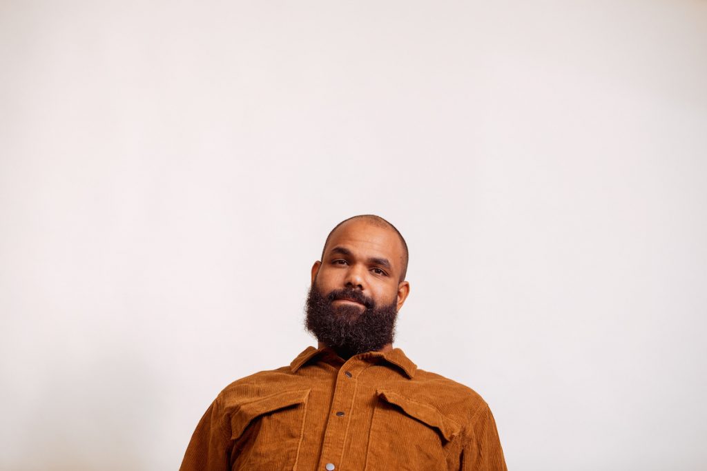 A promotional image of Alan Corduroy Brown. He is wearing a yellow shirt and is against a white wall.