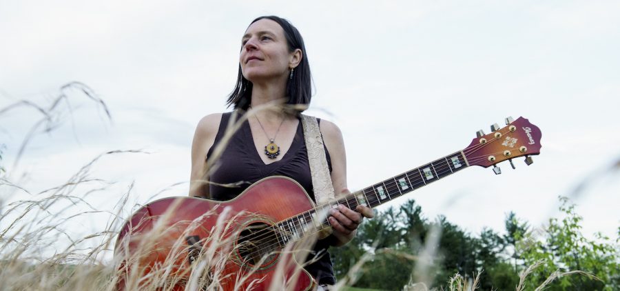A promotional image for singer-songwroter Megan Bee. She is in a field holding a guitar.