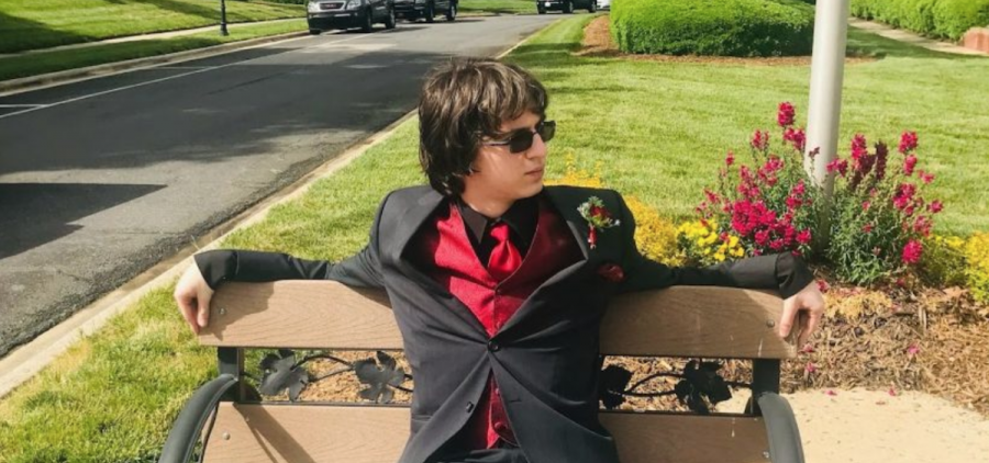 An image of Nicholas Kobe. He is wearing a black suit with ared tie sitting on a bench in a suburban neighborhood.