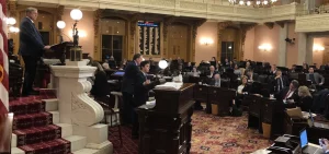 A member of the Ohio Senate gives a speech at the front of the room while other members look on.