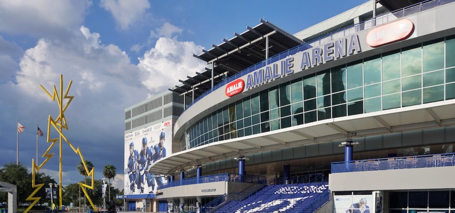 The outside of Amalie Arena in Tampa Florida on a sunny day. You can see glass windows on the upper part of the wall and the Amalie Arena Logo in giant blue letters.