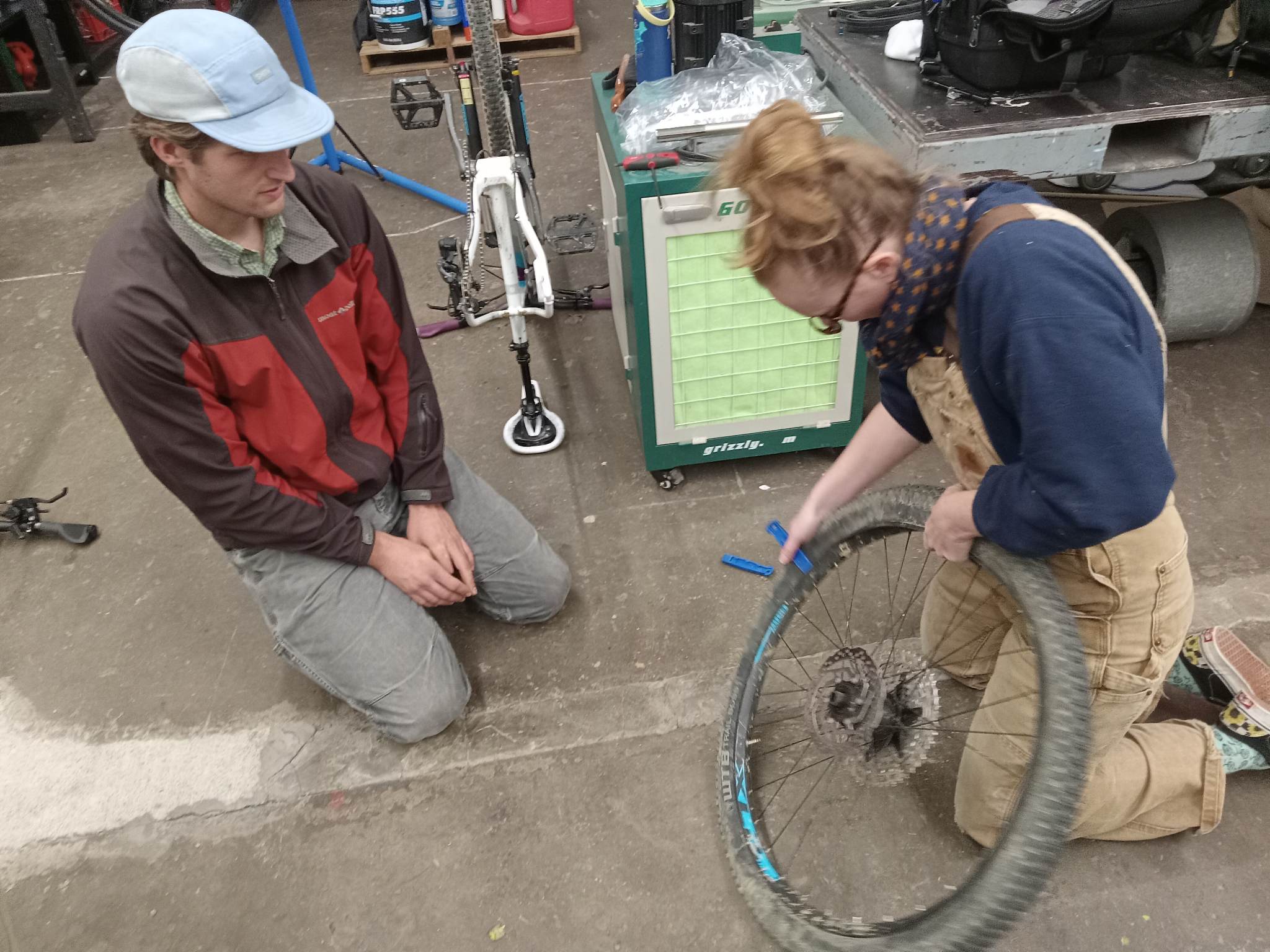 An image of two people kneeling down examining a bike tire, they are on a concrete floor.