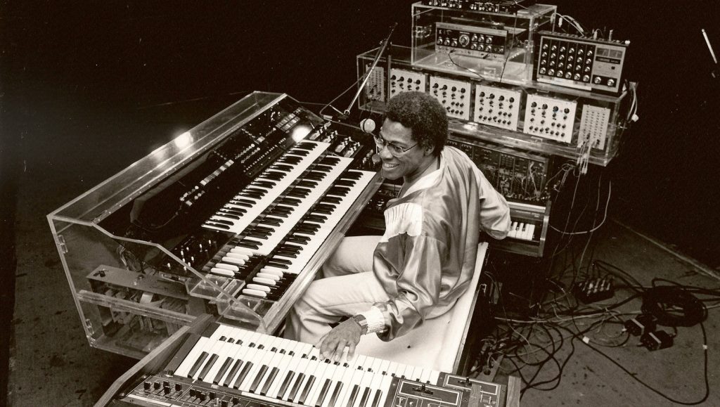 A black and white image of Don Lewis playing his Live Electronic Orchestra, which looks like many synthesizers surrounding him.