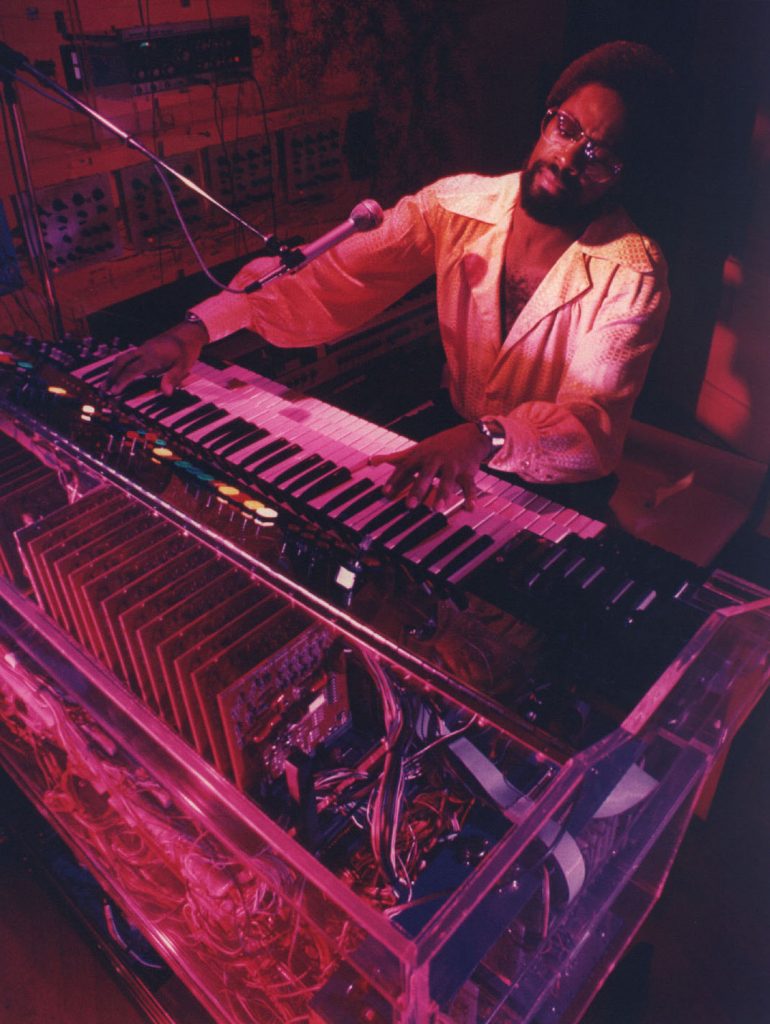 An image of musician Don Lewis, under stage lights that are red. He is playing a synthesizer.
