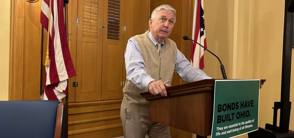 Former Ohio lawmaker and newspaper editor Mike Curtin talks to reporters at the Ohio Statehouse during a press conference. He's at a small podium with a sign on the front that reads Bonds Have Built Ohio. They are essential to the quality of life and well-being of all Ohioans.