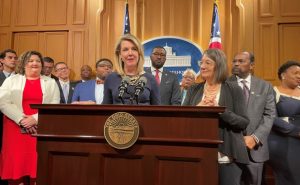 Rep. Allision Russo (D-Upper Arlington), House Democratic leader, stands at a podium with many of her fellow Democratic caucus members in the Ohio Statehouse press briefing room.