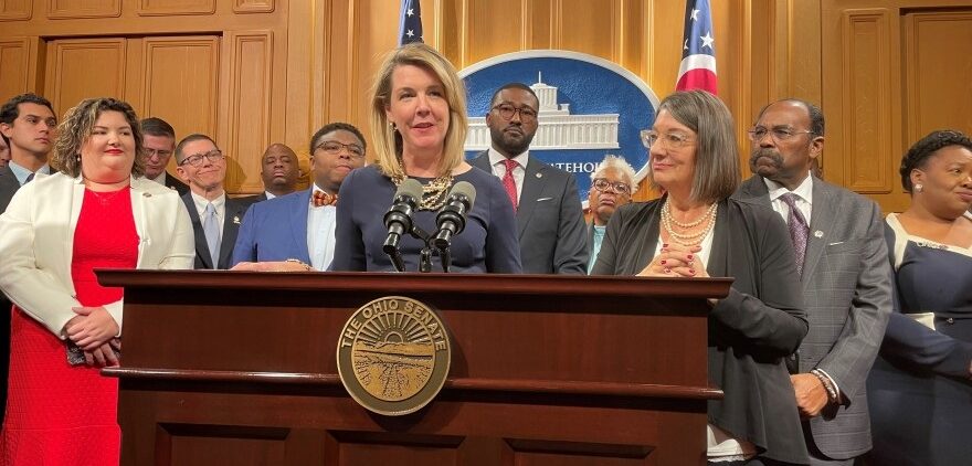 Rep. Allision Russo (D-Upper Arlington), House Democratic leader, stands at a podium with many of her fellow Democratic caucus members in the Ohio Statehouse press briefing room.