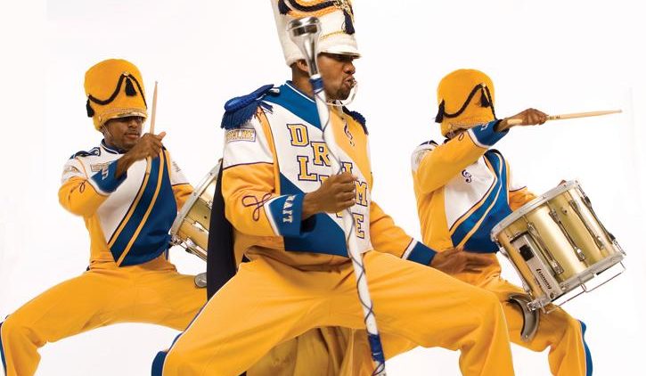 A promotional image for DRUMline, depicting three members of DRUMline in yellow marching band uniforms against a white backdrop.