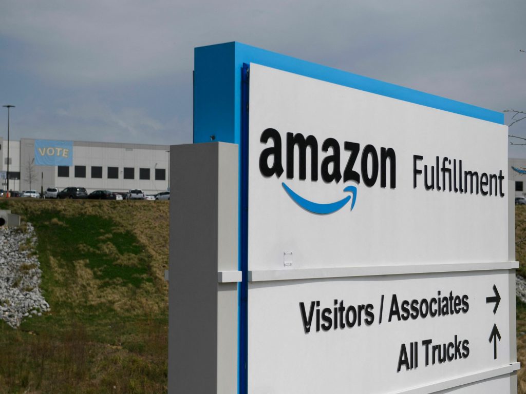 The Amazon fulfillment center in Bessemer, Alabama photographed from just beyond a sign with the building in the back. The building has a vote sign hanging off the side