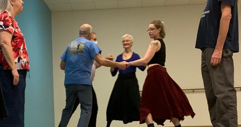 An image of six people in a room dancing. Four of them are holding hands in a circle and two are to the side watching.