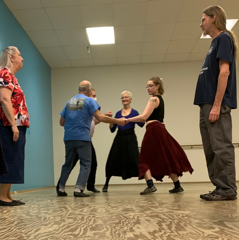 An image of six people in a room dancing. Four of them are holding hands in a circle and two are to the side watching.