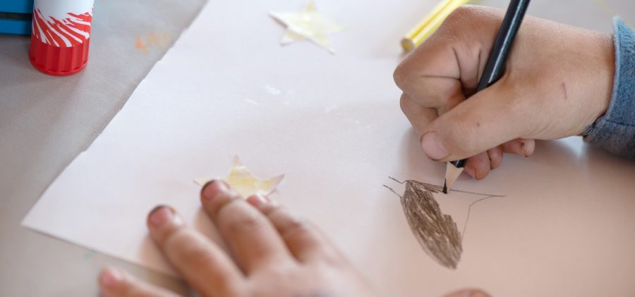 A child's hands draw with a colored pencil on a white piece of paper on a desk.