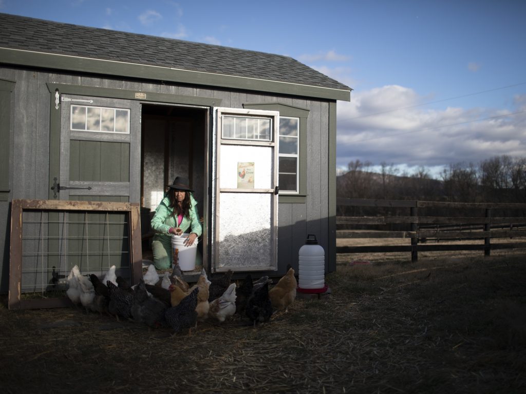 A woman sits on the stoop of a chicken coop, feeding chickens.