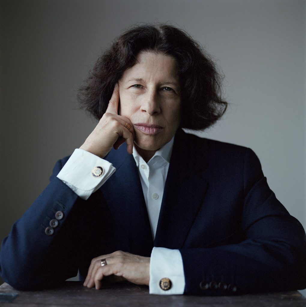 A promotional image of Fran Lebowitz. She is pictured from the waist up, with her elbows on a surface in font of her. She has her right hand against her face, and her left hand on the table. She is wearing a suit.