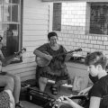 A black and white photo of a group of people engaged in a traditional stringed instrument lesson on a porch.