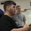 A Jackson High esports team member sits in a gamer chair with a controller in his hand as a fellow team member watches.