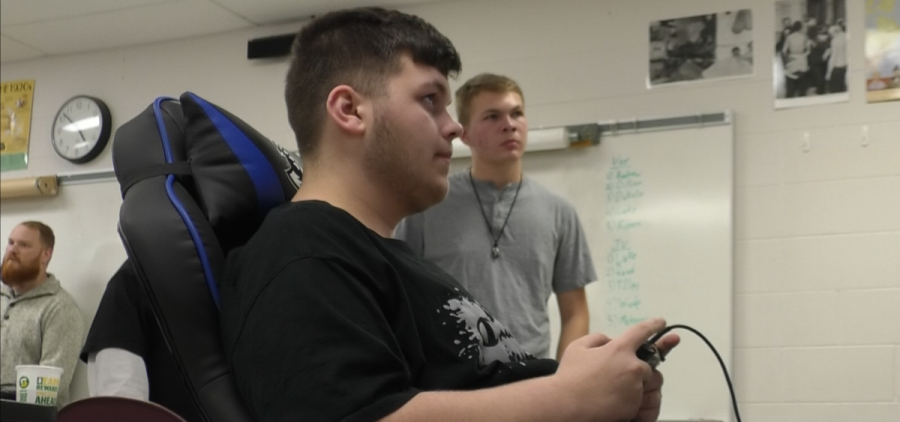 A Jackson High esports team member sits in a gamer chair with a controller in his hand as a fellow team member watches.