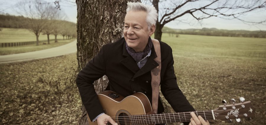 TOMMY EMMANUEL holding guitar leaning against tree in the park