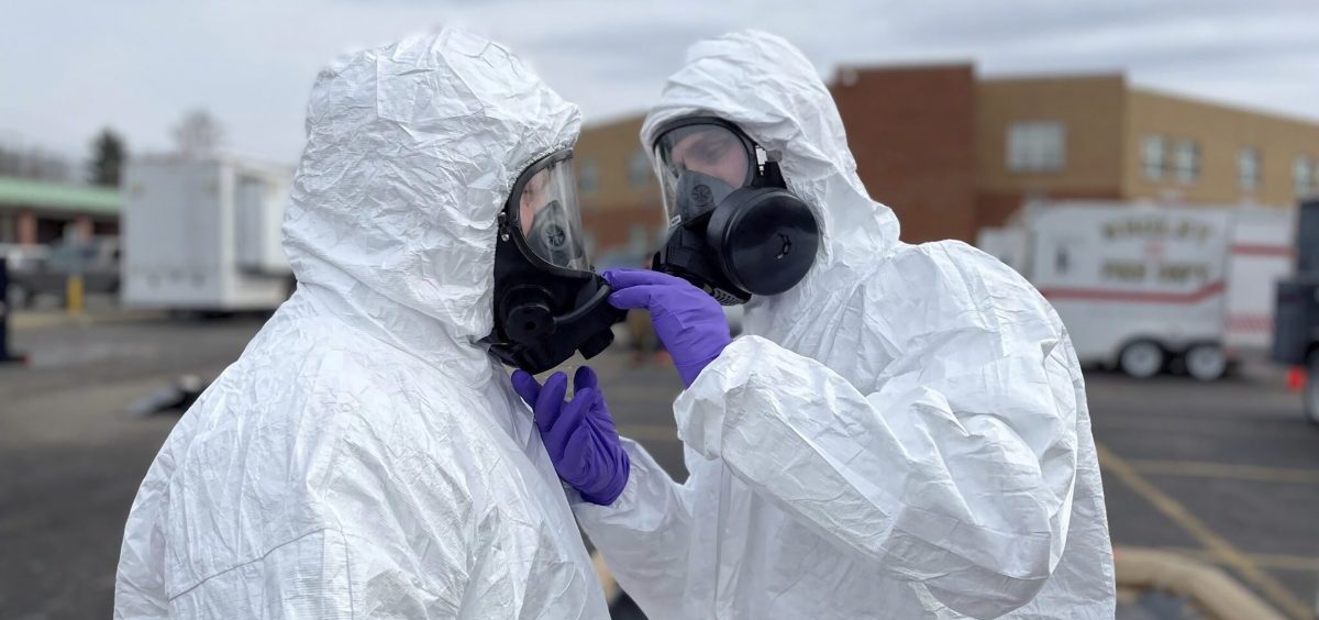 Two individuals wear gas masks and white body suits to protect themselves from hazardous materials.