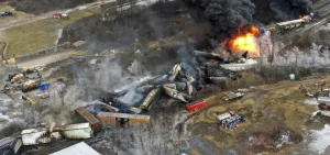 An overhead shot of rail cars shown off the track with some on fire.