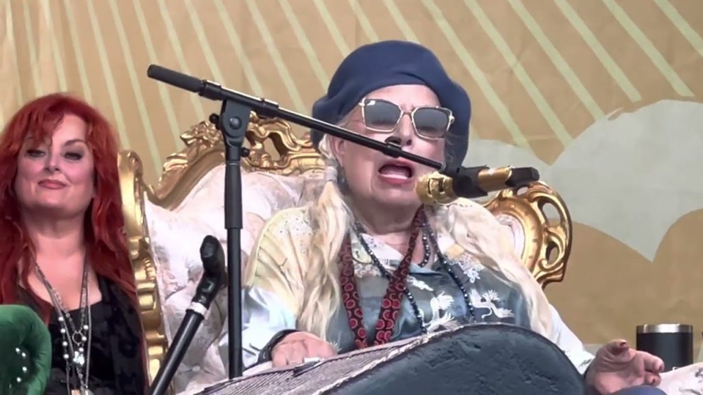Joni Mitchell sings at the 2022 Newport Folk Festival while sitting down. Behind her is Wynonna Judd.