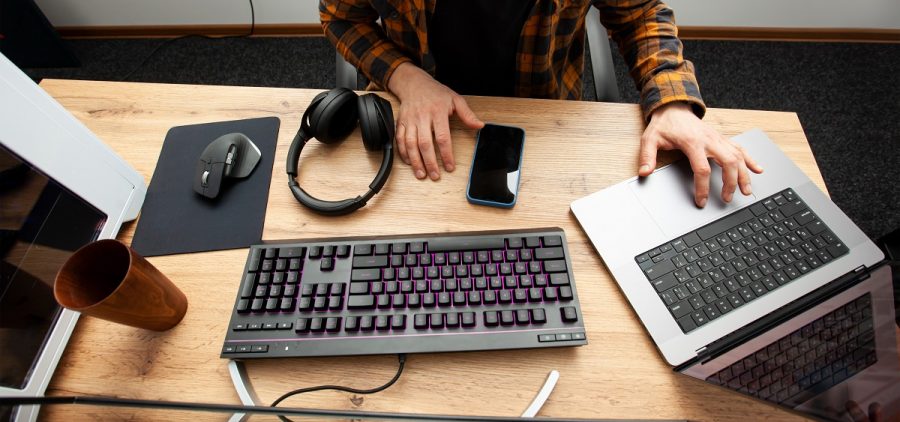 A guy works on a laptop at his desk. He has headphones, a keyboard, a mouse and his phone in front of him.