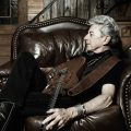 A promotional photo of musician Joe Ely. He is reclining in a chair in a dark room.