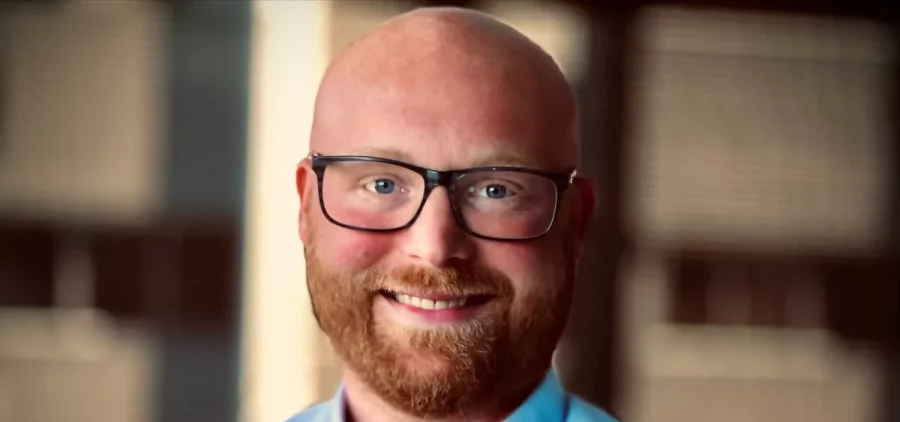 Tyler Fehrman poses for a portrait. He a bald, white man with a red beard and black-framed glasses.