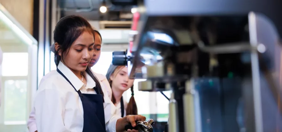 A young girl learns to make coffee in a coffee shop as two other young girls watch behind her
