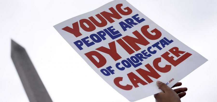 A person holds a white sign that reads Young people are dying of colorectal cancer in red and blue lettering with the washington monument in the background
